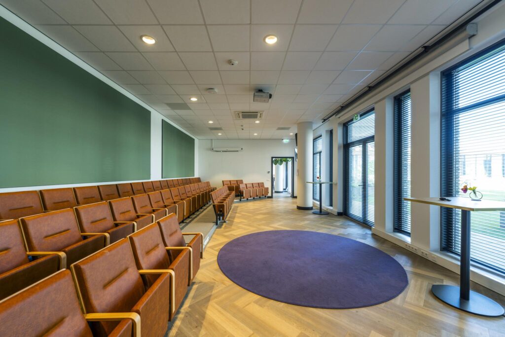 Modern auditorium with brown leather chairs, large windows with blinds, and a purple circular rug on the wooden floor.